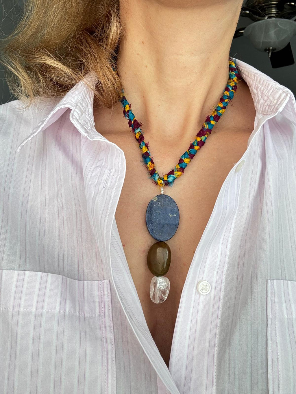 The braided sari silk cord worn at the collarbone, pendant hanging over a white striped shirt