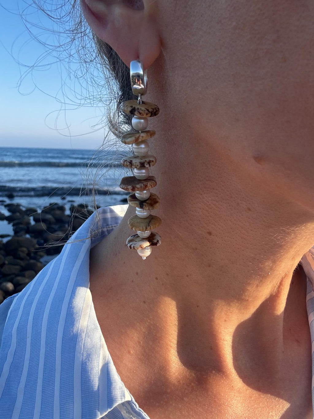 Close view of one earring worn against the sea, showing the brown veining in the magnesite and the polished sterling silver huggie hoop