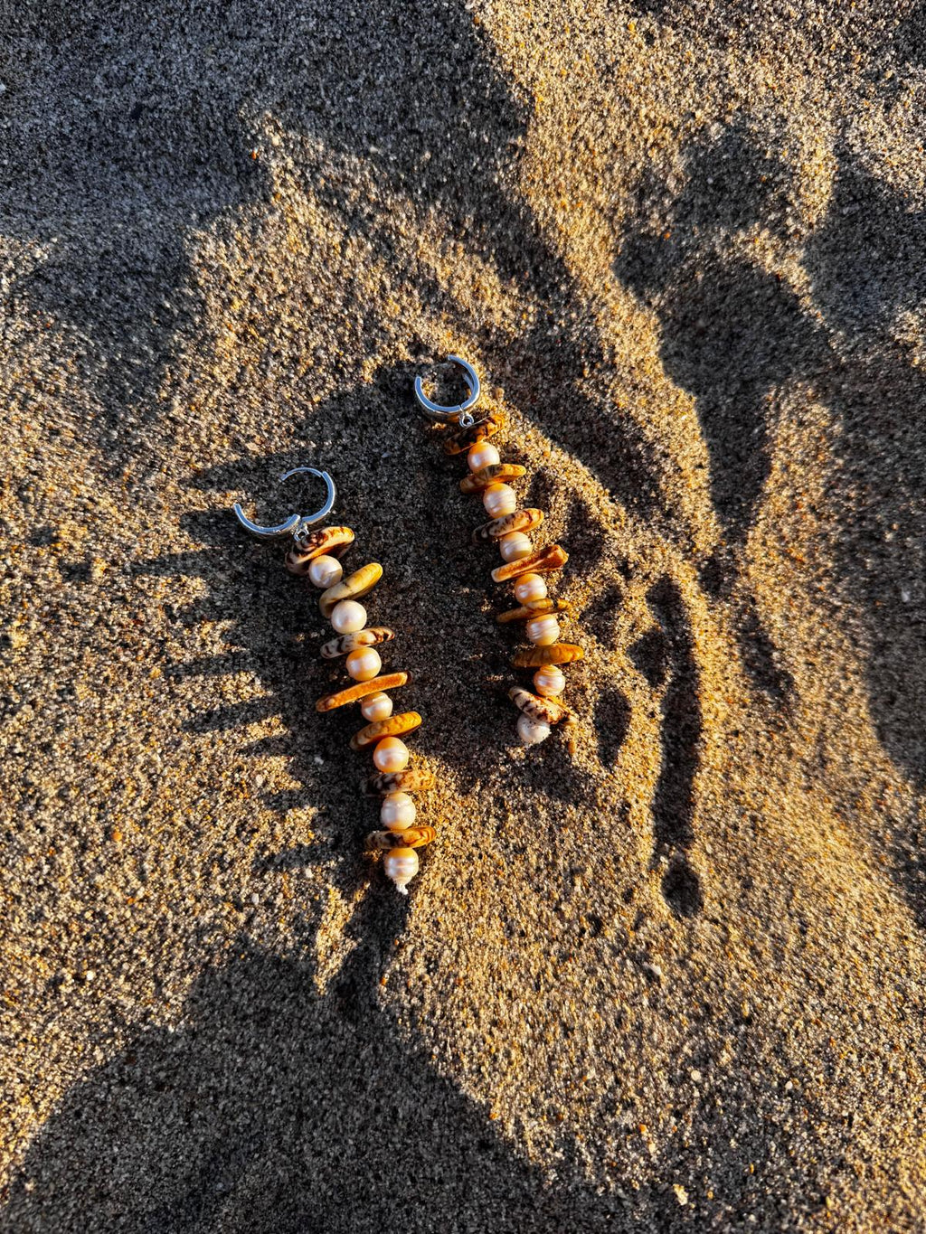 A pair of long drop earrings lying on sand — cream magnesite discs alternating with white freshwater pearls, on sterling silver hoops