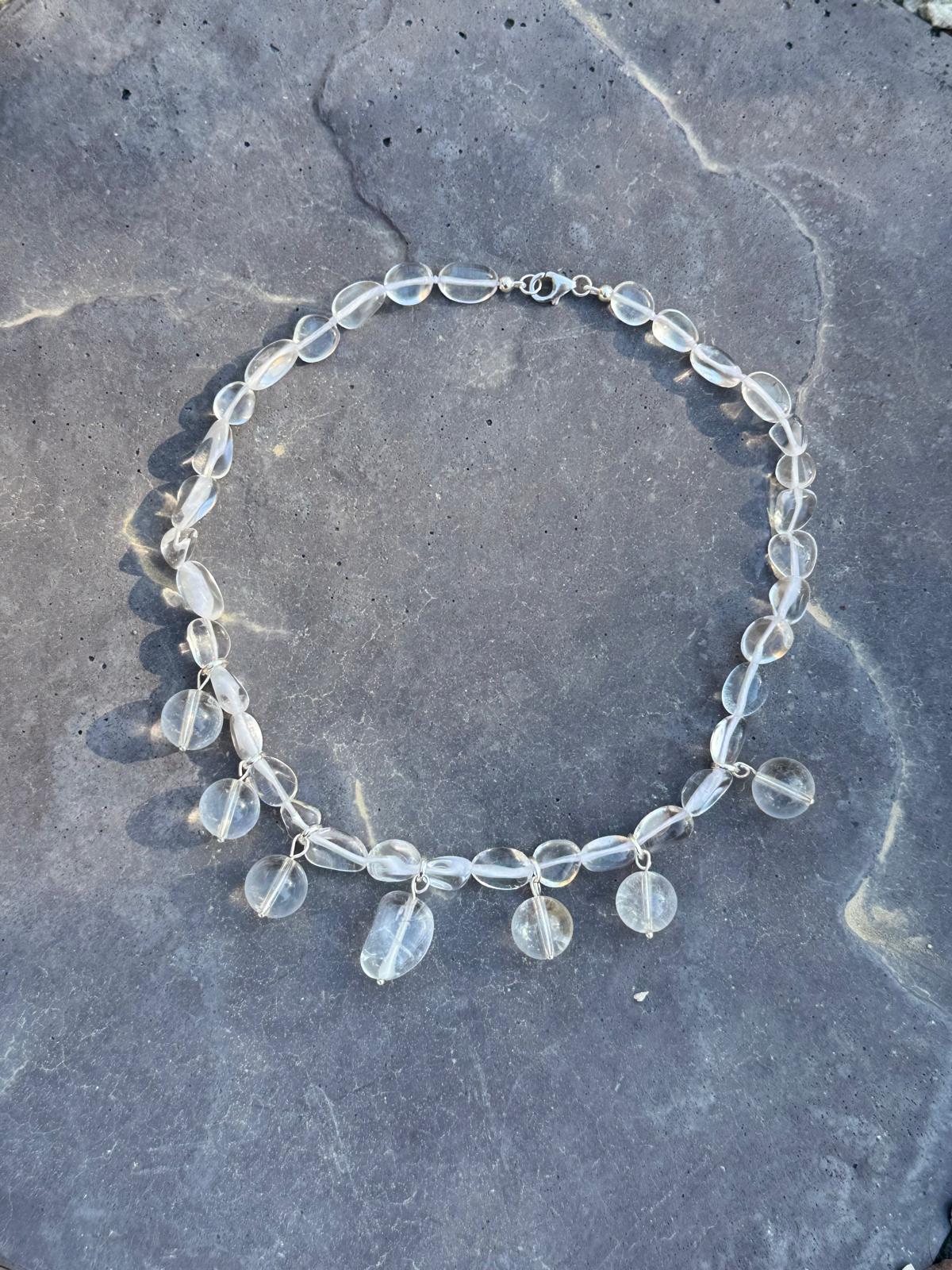 A clear quartz necklace laid on grey stone — tumbled rock crystal beads with seven round drops hung along the front, sterling silver clasp