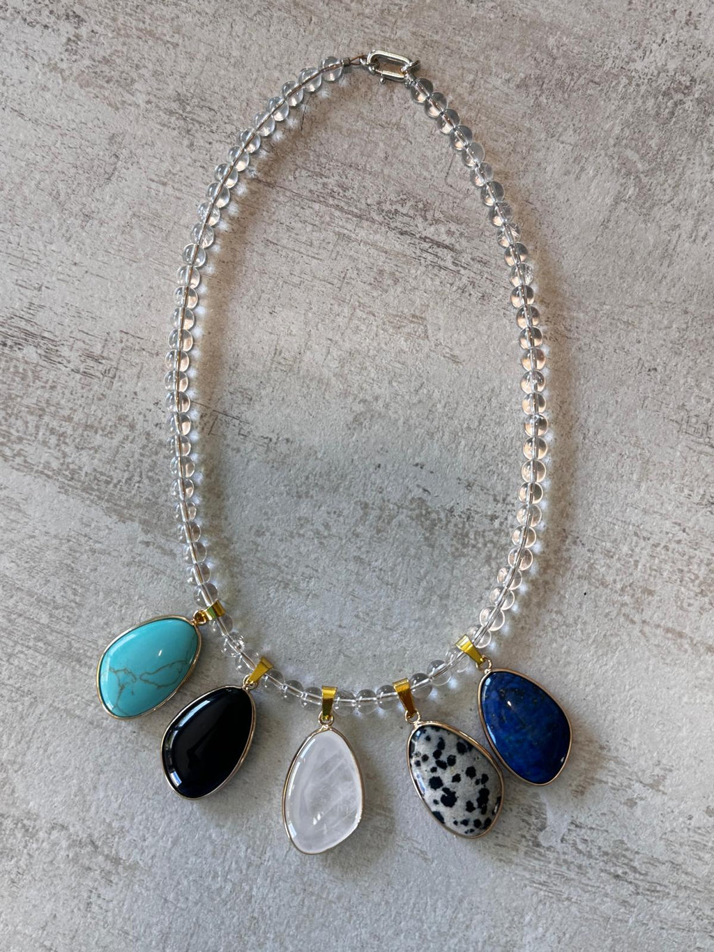 A statement necklace laid flat — five stone cabochons in gold-plated bezels hung along a strand of clear quartz beads