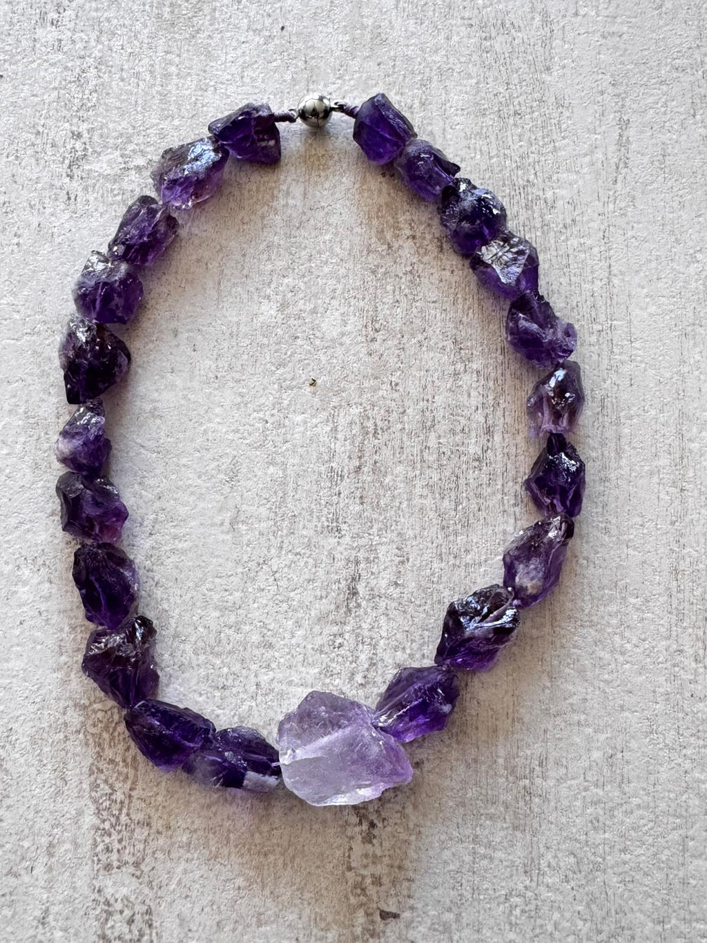 The necklace laid flat — a ring of unpolished deep violet amethyst chunks with one pale stone and a round magnetic clasp