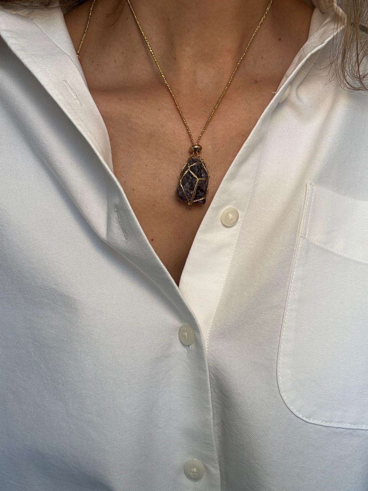 The raw amethyst pendant worn over a white shirt, the stone held in a hand-braided gold net