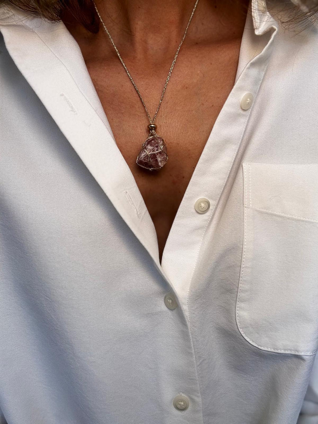 The raw strawberry quartz pendant worn over a white shirt, the pink stone held in a hand-braided gold net