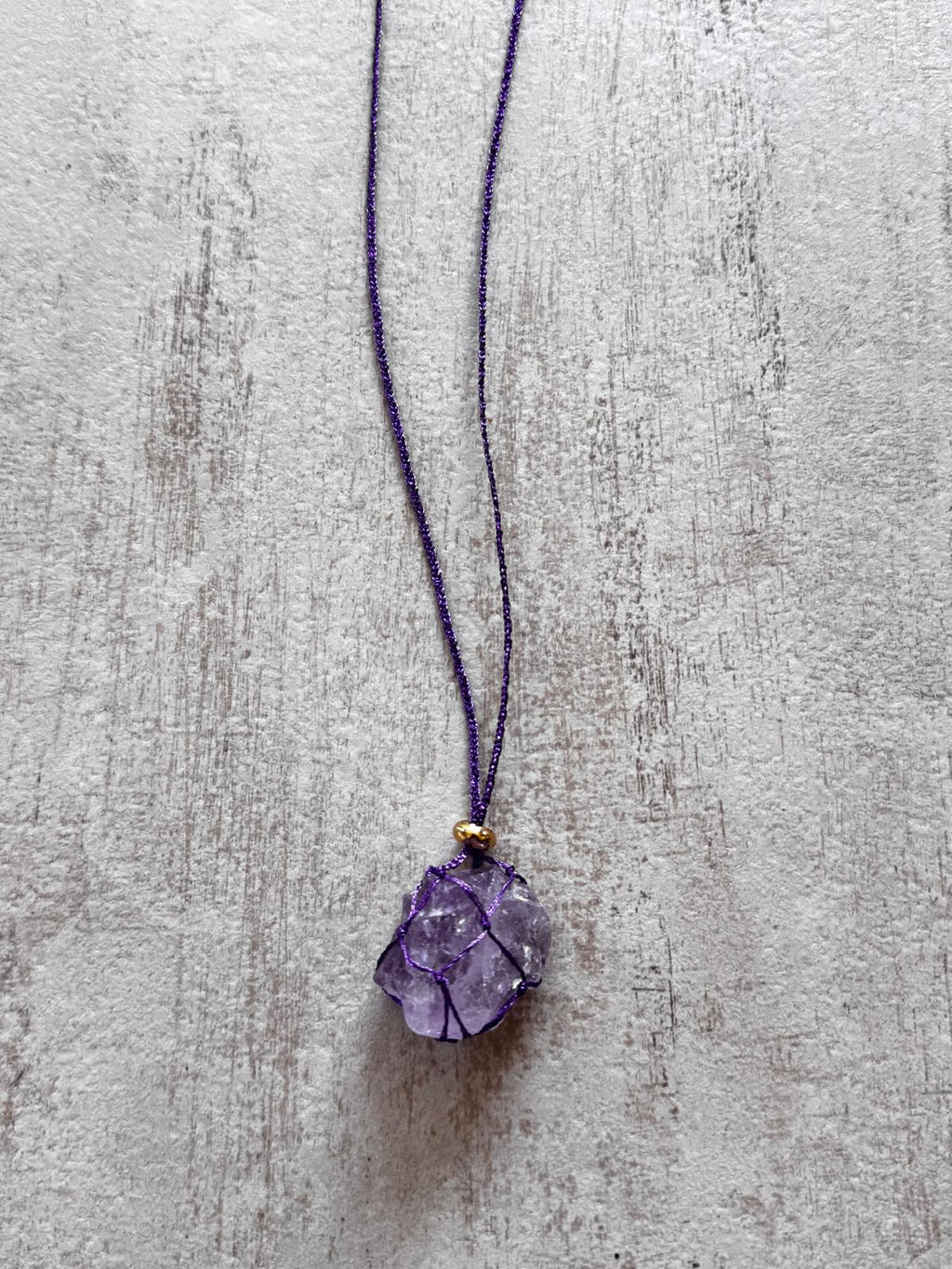 Close up of the raw amethyst pendant held in a purple braided net, gold-plated spacer above